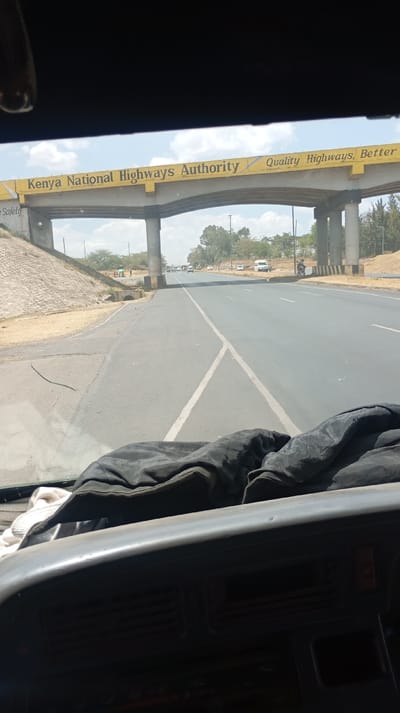 The image captures a view from inside a vehicle, looking out through the windshield onto a highway scene. The foreground shows the dark dashboard of the vehicle, with a dark, crumpled garment (possibly a jacket or blanket) and a white cloth resting on it.

In the midground, a multi-lane asphalt road extends into the distance under a bright, partly cloudy sky. White road markings are visible, including a solid line along the left shoulder and a distinct 'Y' shaped marking in the middle of the road, suggesting a merge or a split. To the left of the road, there is an unpaved shoulder with some sparse vegetation, leading up to a sloped embankment reinforced with grey concrete blocks. A small concrete culvert or drain opening is visible at the base of this embankment.

The most prominent feature in the midground is a large overhead bridge or overpass. Its main beam is painted bright yellow and features dark lettering. On the left side of the bridge, the text "Kenya National Highways Authority" is clearly visible. Further to the right, a slogan continues across the bridge: "Quality Highways, Better Safety". The bridge is supported by concrete pillars on either side of the road.

In the distance along the highway, several vehicles can be seen, including cars and at least one white van. A motorcycle is also visible on the far right side of the road. Trees and bushes line both sides of the highway in the distance. The overall impression is a sunny daytime scene on a well-maintained road.