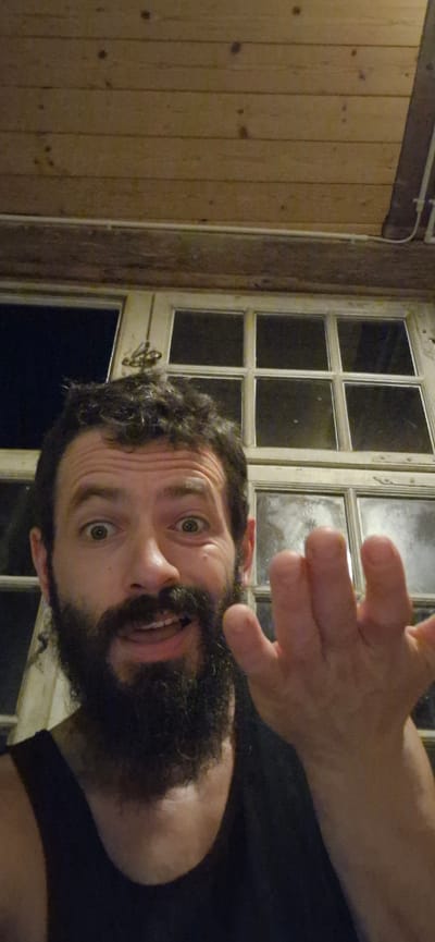 The image shows a close-up, selfie-style portrait of a man with dark curly hair, a full beard, and light brown eyes. He is looking directly at the camera with a surprised or excited expression, his mouth slightly open and his eyes wide. His right hand is raised and extended towards the camera, with his fingers splayed. The man is wearing a dark tank top. Behind him is a large window with multiple panes, appearing to be an older style with a light-colored frame. Outside the window, it is dark, suggesting it is nighttime or an interior space. The background above the window consists of wooden paneling. The scene is indoors. There is no information in the image to suggest a specific location within Wattenwil, Switzerland.
