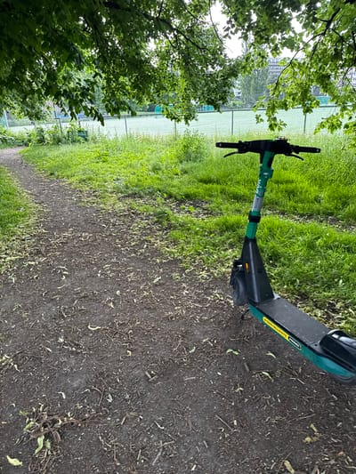 An electric scooter is parked on a dirt path beside a grassy area with a chain-link fence and a sports field in the background. Trees with green leaves overhang the scene, casting shadows. The scooter is green and black with yellow lettering on its deck. The path is covered with fallen leaves and debris. The environment appears to be a park or recreational area, possibly in the city of Oslo, Norway, given the visible text on the scooter that reads "BYENS BILLIGSTE PRISER."