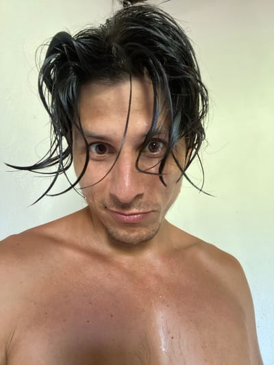 A close-up, chest-up, selfie-style photograph shows a person with dark, wet hair and brown eyes. The person's face is angled slightly towards the camera, with a faint smile. Their skin appears slightly flushed and is dotted with small beads of sweat, particularly on the chest and shoulders. The background is a plain, light-colored wall. The image is taken indoors, possibly in a residential setting in Los Velásquez, Venezuela.