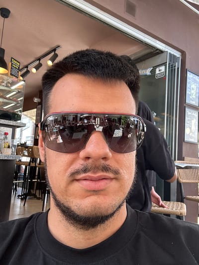 This is a first-person perspective photo of a person wearing large, dark sunglasses. The person has short, dark hair and a goatee. They are wearing a black long-sleeved shirt. The sunglasses reflect a scene with tables and chairs, suggesting the person is likely at an outdoor cafe or restaurant. Behind the person, there are glimpses of wicker chairs and what appears to be a menu board with text. The background also shows a building with large windows and some signage. On a wall to the left, there are a couple of light fixtures and a sign that says "TAPAS 14€". The overall lighting suggests it is daytime, and the scene appears to be casual and relaxed. Given the location context of Marbella, Spain, it's likely a warm and sunny day.