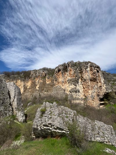 The image shows a rocky cliff face in Kartozhabene, Bulgaria, under a sky with wispy clouds. The cliff is layered with brown and gray rock, and some small trees and shrubs grow along its edges. To the right of the cliff, there are larger gray rocks and some green grass. The sky is blue with long, streaky clouds.