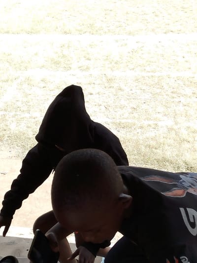 The image displays two individuals outdoors against a bright, light-colored background of sparse ground, likely dry grass or soil. A specific location within Nakuru, Kenya, is not discernible. In the foreground, a person with short, dark hair is observed from the upper back and right profile, leaning forward with their head angled downwards. This individual wears a dark short-sleeved top with a faded, possibly graphic, design on the upper back and shoulder. A dark rectangular object, consistent with a smartphone, is held in their left hand, which is positioned low in the frame. A dark wristband is present on their right wrist. Partially obscured behind and to the left of the foreground subject is a second person. This individual wears a black hooded sweatshirt with the hood pulled up, completely obscuring their face. They are also leaning forward, similarly directed downwards. Both subjects appear to be focused on the lower visual plane, suggesting a shared activity or observation involving the object held by the foreground individual.