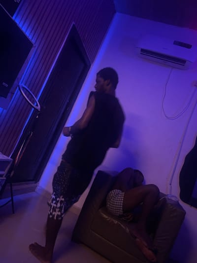 The image depicts an indoor room, likely a living space or studio, bathed in atmospheric blue and purple ambient lighting. The overall scene is dim, suggesting it is night or a room with darkened windows.

Two individuals are present. On the left side of the frame (when viewed upright), one person is captured with significant motion blur, indicating they are moving or in a dynamic pose. They are wearing a dark t-shirt and dark shorts with a distinct white patterned design. A watch or bracelet is visible on their left wrist, and they are barefoot. Their face is indistinct due to the motion blur and low lighting.

On the right side, another person is resting on a dark brown or black armchair/sofa. They are lying on their side with their legs bent, wearing plaid or checkered shorts. Their face is not clearly visible, but their bare feet are prominent.

Key objects in the room include a white wall-mounted air conditioning unit high on the wall on the left. Below it, a dark door is visible, and along the base of the adjacent wall, a blue LED strip provides accent lighting to a textured, striped wall paneling. A ring light on a stand is positioned in the lower-middle section of the image. The floor appears to be tiled or plain light-colored.

The dominant blue and purple color scheme, coupled with the artificial light sources and the ring light, suggests a deliberate aesthetic, possibly for content creation or a relaxed, mood-lit environment. No discernible text is visible. The location context is Ikot Ekpene, Nigeria.
