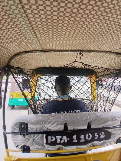 The image provides an interior view from the rear passenger compartment of a three-wheeled auto-rickshaw, looking forward on a street in Kwekwe, Zimbabwe. The vehicle's ceiling is lined with a light-colored fabric featuring a repeating brown checkered pattern. A black mesh cargo net is suspended across the front interior section, above the driver's compartment. A light grey, possibly plastic or fabric, cover drapes over a partition or seat directly in front of the viewer, to which a black rectangular panel displaying "PTA-11052" in white stenciled text is affixed. The back of a male driver, wearing a dark blue shirt, is visible in the front seat. Directly ahead and slightly to the left, another three-wheeled auto-rickshaw is visible through the front opening, featuring a turquoise lower body and a yellow upper body/roof. Its rear license plate, "A-5990L", is partially visible. The background consists of blurred street elements, indicating an urban setting.