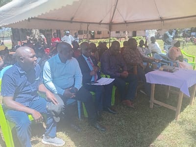 An outdoor gathering is depicted in Idakho South ward, Kenya, under a white tent canopy. Approximately 20-30 individuals, primarily adults, are visible on a grassy area. In the foreground, five men are seated on bright green plastic chairs, facing right. From left to right: a bald man in a dark blue polka-dotted shirt and jeans, holding a dark cap; a bald man in a light blue collared shirt, holding a dark object; a man in a dark suit with a red tie, holding documents; a man in a dark patterned long-sleeved shirt; and another man in a dark patterned long-sleeved shirt. To the right of these individuals, a wooden table covered with a light purple tablecloth holds a black microphone, a dark smartphone, and a small black object. A woman in a light top and patterned skirt is partially visible, seated on a light blue plastic chair further to the right. In the background, more attendees are visible, both seated and standing, with trees and a light-colored building discernible behind them. The individuals in the foreground appear attentive, suggesting observation of an ongoing event or speaker located off-frame to the right.