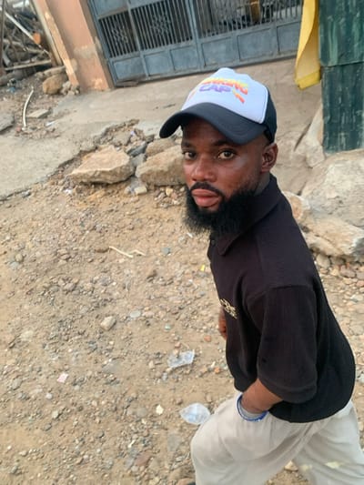 A man wearing a black ribbed shirt, white pants, and a white and black baseball cap with "Viking Cap" printed on it, stands outside on rocky terrain. His beard is dark and full. He is looking directly at the camera with his tongue slightly protruding. In the background, there's a building with a fenced window area, a patch of yellow material, and scattered debris on the ground.