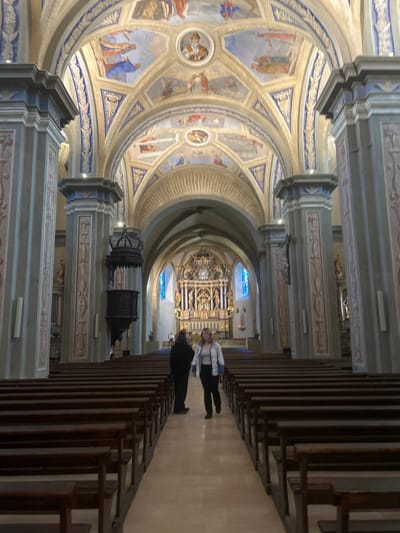 The interior of a grand church features ornate architecture. A frescoed dome with religious scenes overlooks the nave. Rows of wooden pews line the sides of the aisle, leading towards a decorated altar and stained-glass windows. Two figures are walking towards the altar, standing out amidst the intricate details of the church's design. The light illuminates the artistic patterns on the ceiling and walls.