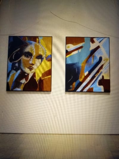 The image displays two framed artworks mounted side-by-side on a light-colored wall in an indoor environment. The specific location within the city of Reifild-Ist, Nigeria, is not identifiable from the visual information provided.
The left painting features a semi-abstract depiction of a human face, possibly female, wearing a dark hat. The color palette includes prominent yellows, golds, browns, blues, and black, with defined lines and shadows contributing to an expressive style. The right painting presents an abstract composition characterized by interconnected geometric and organic shapes, rendered in shades of blue, brown, black, white, and light orange/brown. Both artworks are enclosed in dark, thin frames.
The wall is a plain, light-colored surface, likely off-white or cream, extending above and below the paintings. The floor is a light-toned surface, appearing to be wood or laminate, with visible planks or panels that exhibit subtle reflections. No people or active interactions are visible in the scene. Faint linear artifacts, possibly hairs or scratches, are discernible in the upper section of the image, particularly above the left painting. The overall image exhibits characteristics consistent with a photograph of a digital display, including subtle screen-line patterns on the wall and floor.