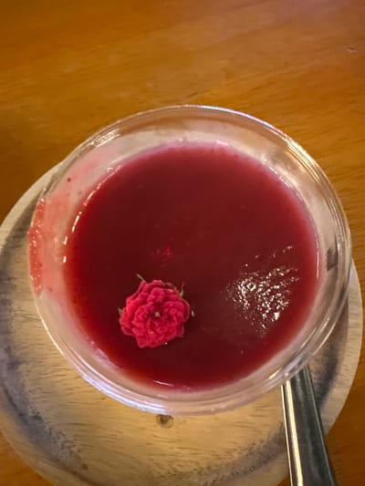 A close-up, overhead shot shows a clear plastic bowl filled with a bright red, smooth liquid, possibly a dessert or drink. A small, vibrant pink rosebud sits on top of the liquid. The bowl rests on a light-colored wooden coaster, which in turn is placed on a textured, reddish-brown wooden surface that appears to be a table. A metallic spoon is partially visible on the right side of the frame. The lighting is soft, highlighting the sheen of the liquid and the delicate petals of the flower. The overall scene suggests a peaceful moment, perhaps at a cafe or restaurant in Patong, Thailand, where one might enjoy a refreshing treat.