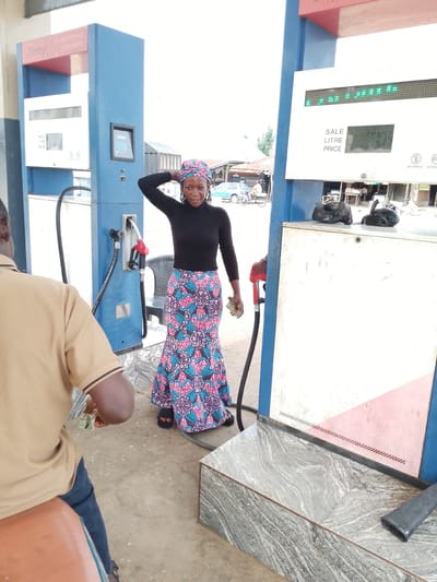 In Serti, Nigeria, a woman stands at a gas station, her hand casually resting on her head, her other hand holding a wad of cash. She is wearing a black turtleneck and a colorful, patterned skirt. The gas pump she is standing next to displays a sale of 500.00, with a litre price of 0.39. The scene takes place outdoors, with a light sky visible in the background. A man in a tan shirt is partially visible in the foreground, facing away from the camera. The gas station appears to be a functional, albeit simple, establishment.