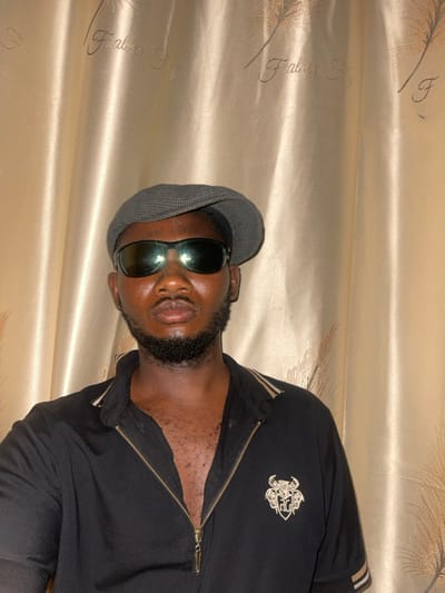 A man in a flat cap and sunglasses poses against a shiny gold curtain. He wears a partially zipped black jacket with a detailed logo on the chest and tan stripes on the sleeves. His beard is neatly trimmed. The angle makes the background appear to be on the side. The lighting casts reflections on the glasses.
