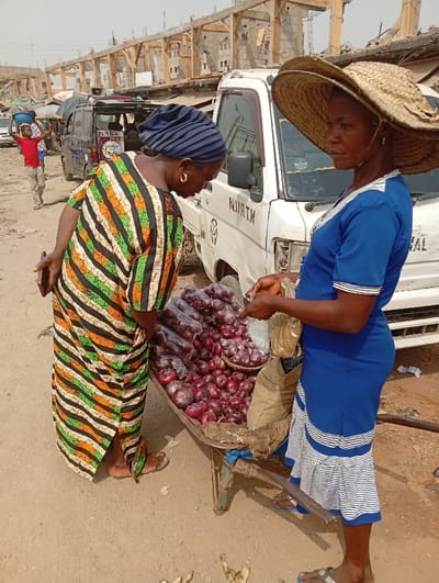 In a bustling marketplace, two African women engage in commerce. One, adorned in a colorful striped dress and a blue headwrap, examines onions displayed on a handcart. The cart is filled with loose and packaged onions. The vendor, wearing a blue dress and a wide-brimmed straw hat, prepares to bag onions for the customer. A white van with lettering on the side stands nearby. In the background, a man carries a container on his head, and buildings under construction add to the busy scene.