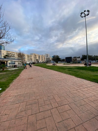 The image depicts a public promenade area at the Front de Mer in Oran, Algeria, under an overcast sky with some lighter patches. A wide, reddish-brown paved walkway, featuring an irregular block pattern, extends from the foreground into the mid-ground, gently curving to the right. Green grass borders both sides of the walkway.
On the left side of the scene, a series of multi-story buildings with balconies and visible air conditioning units line the background. The building closest on the left features signage that reads "CAPRI CAFÉ "GAMBROUX"". A taller, modern building with a blue facade is partially visible behind it. Bare tree branches from a deciduous tree are present in the far left foreground.
In the mid-ground, several individuals are present. On the paved walkway, a group of at least three people stands together on the left side. Further down the path, more individuals are visible. In the grassy area to the right, multiple people are seated on white benches; one group consists of at least three individuals, and another group of two.
A tall, grey metal streetlight with a circular array of multiple LED floodlights is prominently positioned on the right side of the mid-ground. Further in the background on the right, a large, dark rectangular structure, resembling an outdoor screen or billboard, is visible within a grassy expanse. The overall environment suggests a leisure area within an urban setting.