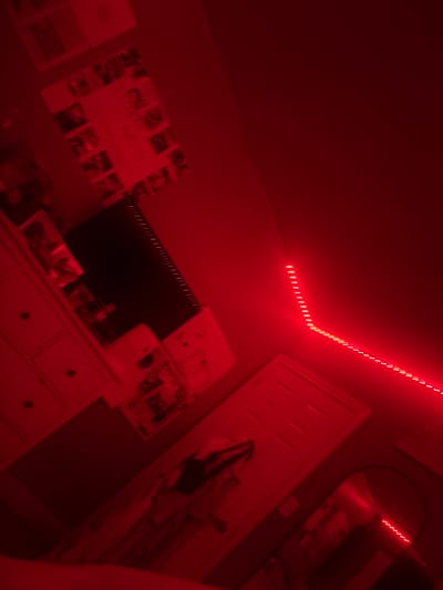 The image depicts a room illuminated by red LED strip lights, casting a strong red hue over the scene. The room contains a white dresser with drawers, a television screen, and posters or pictures affixed to the wall. A closed white door is visible, along with a mirror. The overall environment suggests a teenager's bedroom. The location in Whitby, Canada, is not discernible from the visual information provided.