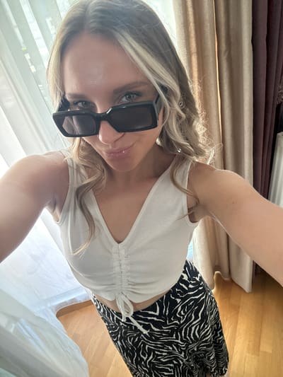 A blonde woman wearing black rectangular sunglasses, a white ruched tank top, and a black and white zebra-print skirt is shown in a selfie. She is indoors, with sheer white curtains and brown curtains visible in the background. The flooring appears to be polished wood. The image was taken in Esenyurt, Turkey.