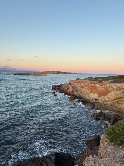 This is a vertical, eye-level shot of a rocky coastline with the sea and a sunset sky. In the foreground, dark, jagged rocks with some green shrubbery descend towards the dark blue sea, which has white foam lapping against the shore. The middle ground features a sheer cliff face of reddish-orange rock, with evidence of stratified layers and erosion. Further out, the sea stretches towards a chain of islands and a distant mountainous coastline, illuminated by the soft hues of a sunset. The sky is a gradient of pale blue at the top, transitioning to pink and orange near the horizon, with a few wispy clouds. The scene is set at the coast of Limenas Markopoulou, Greece, during the evening. There are no people or animals present.