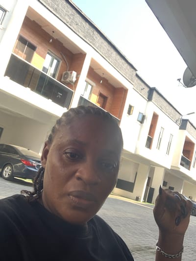 The image depicts an individual positioned in the foreground of a modern building complex located in Ikota, Nigeria. The subject, a person with dark skin and braided hair, occupies the lower half of the frame, looking directly at the camera with a neutral expression. They are wearing a dark-colored shirt. A silver-colored, chain-link bracelet is visible on their left wrist, and the fingernails of their left hand are painted black.
The background consists of at least three multi-story buildings with contemporary architecture, featuring white facades complemented by light brown, possibly wooden, paneling. Architectural details include glass balconies or railings and visible external air conditioning units. A dark-colored sedan is parked on the interlocking concrete block pavement to the left of the subject. A satellite dish is mounted on a pole against the clear, bright sky on the far right. The entire scene is brightly illuminated by natural daylight.