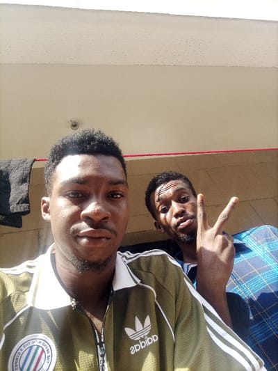 This image depicts two males in an outdoor urban or residential setting, identified as Nibo, Nigeria.
The subject in the foreground, positioned to the left, is a male with dark, short curly hair and a light beard. He wears an olive green track jacket with white stripes on the sleeves and a white collar, featuring an "adidas" logo and a circular emblem on the chest. He faces forward, looking directly at the camera with a neutral expression.
The second subject, positioned behind and to the right of the first, is a male with short dark hair and a sparse beard. He wears a blue plaid long-sleeved shirt. He is looking towards the camera with a slight upward tilt of his head and is making a V-sign (peace sign) with his right hand, fingers extended.
The background consists of a light-colored building exterior. An overhead light-beige ceiling or soffit is visible, with a small, round, light-colored object (possibly a light fixture or small camera) attached to it. A red rope or cord extends horizontally across the mid-background. To the far left, dark fabric, possibly clothing, is seen hanging over a dark railing or line. Sunlight is present, casting highlights on the subjects and illuminating the scene from above and slightly to the right.