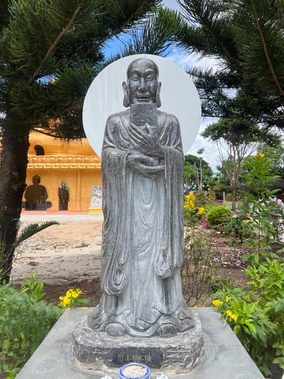 A tall, grey stone statue of a Buddhist monk, identified as "TÔ A-NAN-ĐA" on its pedestal, stands centrally in an outdoor setting within the city of Đăk Mar, Vietnam. The monk statue has a serene expression, elongated earlobes, and holds a rectangular object with both hands in front of its chest. A large, white circular disc serves as a backdrop behind the monk's head. To the statue's left, a multi-tiered, golden-yellow temple building with ornate architectural details and arched openings is visible; a dark silhouette of a seated Buddha statue is discernible within one archway. The ground in front of the temple building is unpaved earth. To the statue's right and in the mid-background, a landscaped garden features various green shrubs, trees, and flowering plants with yellow and pink blossoms. The upper quadrants of the image are partially framed by green pine tree branches. The sky above is partly cloudy blue. At the base of the statue's pedestal, a small blue and white ceramic bowl containing ash or sand, along with two smaller white objects, is present.