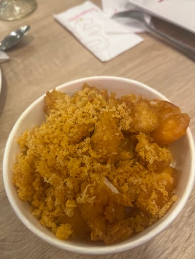 A close-up shot shows a white bowl filled with fried food, possibly chicken or fish, covered in a golden-brown crispy coating. The food appears to be served on a wooden table, with a white napkin and a spoon visible in the blurred background. This image was taken at a restaurant in South Tangerang, Indonesia.