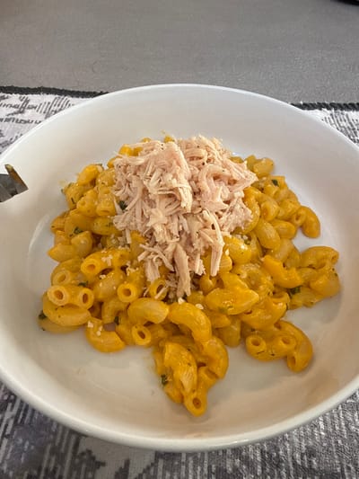 A white bowl holds a portion of macaroni and cheese, mixed with what appears to be herbs. Atop the pasta sits a mound of shredded, pale chicken. The dish is positioned on a surface that features a patterned rug. The angle is a high-angle shot, providing a clear view of the meal's composition.