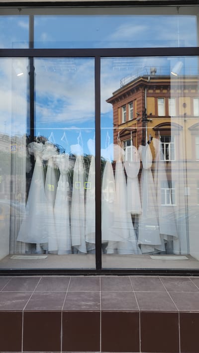 The image shows a shop window, likely a bridal or formal wear store, in Smolensk, Russia. The window displays several white gowns covered in protective plastic. The reflection in the glass shows a building across the street with brown and yellow brickwork, indicating a European architectural style. The sky's reflection suggests a sunny day with some clouds. The right edge of the image captures the exterior wall of the building, which is tiled with brown and grey squares.