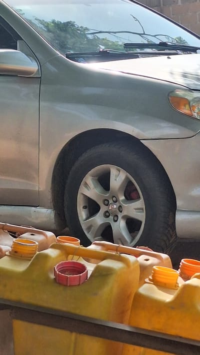 The image shows the side of a silver vehicle, likely a van or SUV, with its tire and wheel clearly visible. The tire is black and appears to be in good condition. The wheel is silver and has a multi-spoke design. The vehicle is parked next to a bright yellow object, which appears to be a large plastic container with multiple orange spouts or openings and a red cap. The background is out of focus, but a brick wall is partially visible on the left side of the image. Sunlight is reflecting off the vehicle's surface, suggesting it is daytime. The overall scene is outdoors, with some greenery reflected in the vehicle's window, indicating a natural setting. There is no visible text in the image. The orientation of the image is such that the vehicle is positioned vertically.