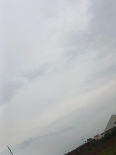 The image captures an overcast sky with scattered clouds, suggesting a diffused light or pre-rain conditions. A power pole with wires is visible in the lower left quadrant, angled upwards. The middle ground shows a line of buildings and vegetation. To the right, a large, light-colored, angular structure dominates the skyline, possibly a modern warehouse or industrial building. The ground appears to be a mix of reddish-brown soil and green grass. The scene is situated in Rafin Bauna, Nigeria, indicating a semi-urban or peri-urban landscape. There are no people or discernible activities in the image.