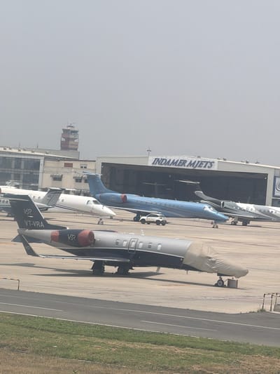The image shows an aerial view of an airport tarmac in New Delhi, India. Multiple private jets are parked, with one in the foreground covered with a protective tarp. In the background, a control tower and a hangar with "INDAMER MJETS" written on it are visible. The sky is overcast.