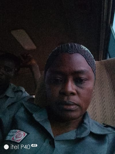A medium shot captures a dark-skinned individual with a netted head covering, possibly in a vehicle. The person wears a uniform with a visible badge on the left chest. Another individual, partially visible, sits in the background to the left, also wearing a similar uniform. The lighting is dim, with an overhead light fixture visible. The image seems to have been taken with an Itel P40 phone, with the watermark present in the lower-left corner. The overall setting is indoors, likely inside a bus or car, based on the seat and window visible.