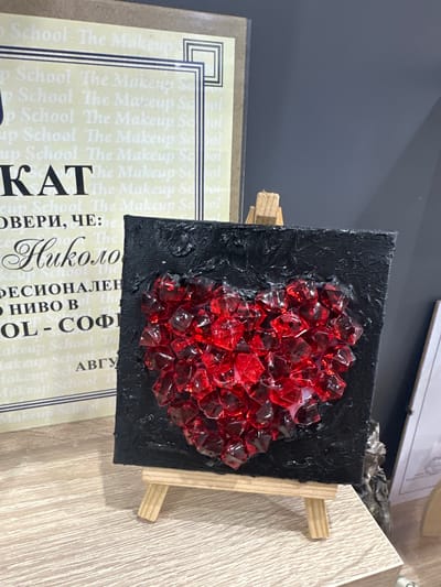 This media file shows a close-up of a small, black, textured canvas art piece adorned with a cluster of red, faceted crystals, resembling a heart or a geode. The artwork is propped up on a miniature wooden easel, with two light-colored wooden supports visible.
In the background, a sheet of paper with repeating text in a light color is partially visible. The text appears to be "School The Makeup" repeated multiple times, with some larger, bolder text in Cyrillic characters, including "КАТ", "ОВЕРИ, ЧЕ:", "Николо", "ФЕСИОНАЛЕН", "• НИВО В", "OL - СОФІ", and "АВГУ". This suggests a potential connection to a makeup school or academy, possibly advertising or displaying credentials.
The setting appears to be indoors, likely a desk or a shelf, with a wooden surface in the foreground. The lighting suggests it could be daytime, though no specific time of day is indicated. The overall mood is artistic and decorative, with the vibrant red crystals providing a striking contrast to the dark canvas. No people are visible in the frame. The image's focus is on the art piece and the accompanying text, hinting at its purpose or origin.
