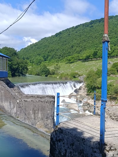 This image depicts a picturesque view of a hydroelectric power station dam situated in Gachedili, Georgia. The dam, constructed of concrete, features a cascading waterfall of clear, flowing water. Adjacent to the dam, a bright blue building with large windows provides a stark contrast to the natural landscape. The surrounding environment is characterized by lush green hills, dense forests, and a vibrant blue sky dotted with white clouds. A metal railing with blue posts and rusted wires lines the walkway, offering a safety barrier. In the foreground, a weathered metal pole, partially painted red and blue, stands tall. A dark electrical wire stretches across the top left of the frame, suggesting the presence of modern infrastructure within this natural setting. The scene is bathed in bright daylight, casting distinct shadows on the concrete surfaces, indicating a sunny day.