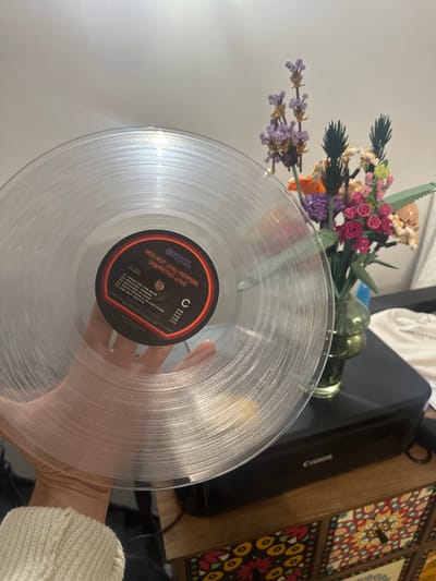 A clear vinyl record, displaying the "Unlimited Love" album by the Red Hot Chili Peppers, is partially held by a hand wearing a white sweater. The record label is visible, listing track titles such as "These Are the Ways" and "One Way Traffic." A bouquet of artificial flowers sits in the background, along with a black printer labeled "Canon" and a decorative tile coaster. The background appears to be a cluttered desk or table.