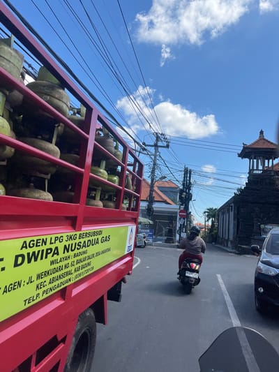 The image captures a street scene in Denpasar Barat, Indonesia, dominated by a red truck carrying numerous yellow propane gas cylinders. The truck is partially visible on the left side of the frame, with its side displaying text in yellow, including "AGEN LPG 3KG BERSUBSIDI DWIPA NUSADUA GAS", an address "JL. WERKUDARA NO. 62, BANJAR DALEM, DESA ANGKANTANA", and a telephone number "0361720139". The truck is positioned on a paved road, indicating it is likely in motion or parked.
In the middle ground, a person is riding a scooter in the same direction as the truck. The scooter's license plate is visible, reading "DK 2387 ABX". The rider is wearing a light brown hoodie and dark pants, with a helmet on. The scooter is in the lane of traffic, and a white line marks the lane division on the road.
The background reveals a street lined with buildings, some with tiled roofs. A distinctive traditional Balinese structure with a multi-tiered roof and intricate carvings is visible on the right side of the image. Overhead, a dense network of electrical wires crisscrosses the sky, typical of urban environments. The sky is blue with scattered white clouds, suggesting it is daytime. A black car is parked on the right side of the road, partially obscuring the view of the building. The overall scene depicts a busy street with transportation and commerce.