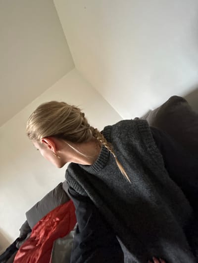 A photograph shows a person with fair hair styled in a braid. The person is wearing a gray sweater. The background consists of a plain, light-colored wall, likely indoors. No details indicate the location is Lyon, France.