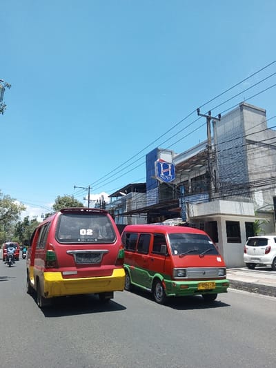 This image depicts a street scene under a clear blue sky, identified as being in the city of Tagtse Tashidingkha, Bhutan. The environment suggests a bright, sunny day.
In the foreground, two distinct public transport vans are prominently featured on a paved road. To the left, the rear of a red and yellow minivan is visible. This vehicle has a red body and a yellow lower rear bumper section. A white "02" sticker is affixed to its rear window, which also shows some internal reflections. The license plate area is empty. To its right, slightly ahead and viewed from the front-right, is another minivan with a two-tone red and green paint scheme. This vehicle has a yellow license plate displaying the alphanumeric sequence "F 4064 ZL" on its front bumper. Both vans appear to be older models, typical of local public transportation in Southeast Asia.
Further down the road on the left, several motorcycles and other vehicles are visible, moving in the same direction. On the right side of the image, alongside the road, a sidewalk or shoulder is present, where a white compact car is parked. Behind the parked car, a multi-story building with a light-colored, possibly concrete, facade is visible. A prominent blue rectangular sign with a white "H" emblem and the text "H HEART" is affixed to the front of this building. Numerous overhead electrical wires are strung across power poles and in front of the building, indicating urban infrastructure. Below the main building and to the right, a smaller, white structure with dark window panes is present at ground level. No specific individuals are clearly identifiable, but the presence of vehicles indicates human activity.