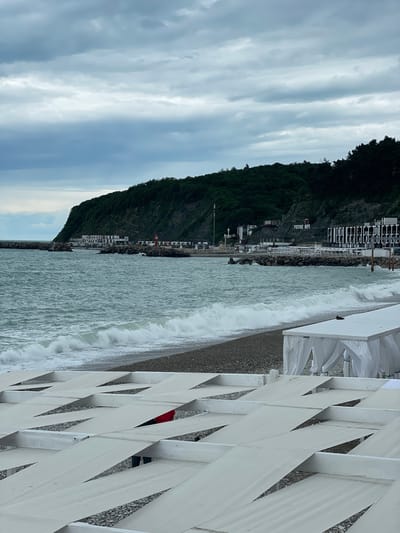 The image captures a coastal scene in Olginka, Russia. The sky is overcast with striated clouds. A steep, forested hillside borders the beach, upon which are buildings and structures. The lettering "Русское Море" is visible on the hillside. Waves from the turbulent sea crash against the shore. A white, lattice-patterned barrier or fence dominates the foreground, with a hint of a red garment or item visible through the lattice. The image is presented on its side.