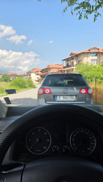 The image depicts a first-person view from the driver's seat of a stationary vehicle, looking forward through the windshield on a clear, sunny day in the city of Bansko, Bulgaria. In the immediate foreground, the top section of a black steering wheel, partially covered with a carbon fiber pattern wrap, is visible. The instrument cluster behind the steering wheel shows a tachometer (left) and speedometer (right), both indicating zero, along with gauges for engine temperature (cold) and fuel level (nearly full).
Directly ahead on an asphalt road, a grey Volkswagen Passat Variant (wagon) is stationary. Its rear displays a Bulgarian license plate with the registration "E 9067 HA". The road appears to ascend gently and curves slightly to the left in the distance. On either side of the road, green foliage, including tall grass and bushes, is present. A brown wire fence runs along the right side of the road.
In the midground and background, multiple multi-story residential buildings with distinctive terracotta-tiled roofs and light-colored facades are visible, characteristic of resort architecture found in Bansko. These buildings feature balconies and some have external air conditioning units. Green trees and dense vegetation are interspersed among the structures. The background reveals clear blue sky with scattered white cumulus clouds, and distant mountains are faintly visible on the far left. No people are visible in the scene. Green tree branches are partially visible at the very top of the frame, arching over the vehicle.