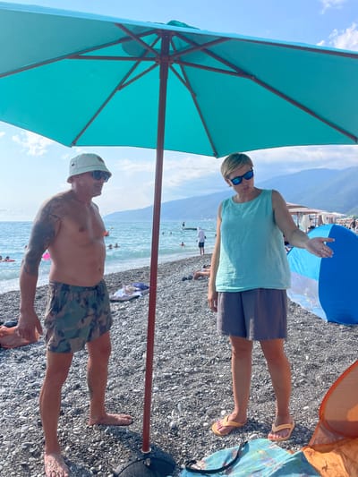 This media file captures two individuals on a pebbly beach in Gagra, Abkhazia. A large turquoise umbrella shades one person, a man wearing a light-colored bucket hat, sunglasses, and camouflage swim trunks. He has tattoos on his left arm. The other individual, standing under a blue and orange umbrella, is a person wearing sunglasses and a light blue top with dark shorts. Their arms are raised as if waving or gesturing. The sea is visible in the background, with several people in the water. The sky is bright blue with some clouds, suggesting it is daytime with good weather. The overall scene conveys a relaxed beach atmosphere, likely during a vacation or outing. There is no visible text in the media file.