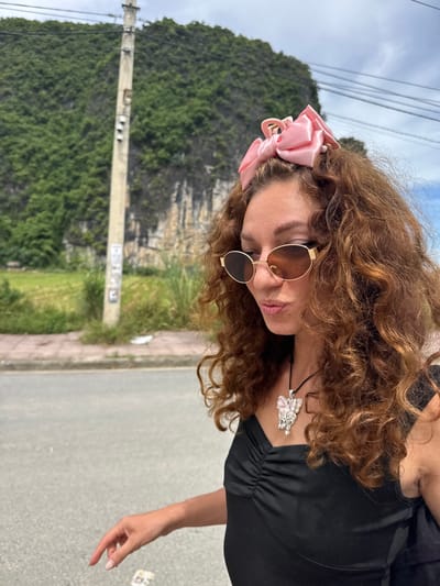 A young woman with curly brown hair is pictured in Nam Hoa Lư Ward, Vietnam. She is wearing a black dress, sunglasses, and a pink bow in her hair. She is also wearing a necklace with a butterfly pendant. The woman is standing in front of a telephone pole and a lush green mountain. The background also shows a sidewalk and a road. The sky is cloudy.