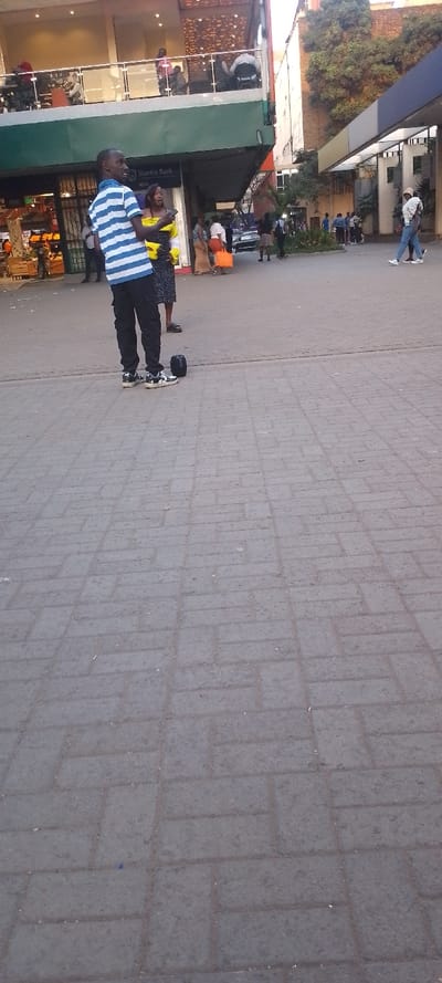 The image captures an outdoor urban scene, likely a public square or pedestrian area in front of commercial buildings. The ground is paved with rectangular grey and dark grey interlocking bricks, showing some signs of wear and dirt.
In the foreground, a man stands facing slightly to the right, looking forward with a slight smile or smirk. He is wearing a blue and white horizontal striped short-sleeved shirt, black cargo pants, and black and white sneakers. To his right, near his feet, is a black object that resembles a single wheel of a hoverboard or a unicycle-like personal transporter.
Behind the man, a crowd of people is visible. To the man's left and slightly behind him, a woman with long dark hair, wearing a yellow top and a dark patterned skirt, is looking towards the left, holding a phone to her ear or hand. Further into the midground, several other individuals and small groups of people are standing or walking. Some are carrying bags; one person on the right side of the midground carries an orange bag. In the background, what appears to be a street with parked vehicles can be seen, and more pedestrians are present.
The buildings on the left side of the image are multi-story commercial structures. The lower level has large windows, and signage for "Stanbic Bank" is clearly visible. Above this, there's a green canopy or awning, and a second story features an open balcony or terrace with glass railings, where more people are visible, some seated. To the right of these buildings, there's a denser urban environment with trees and other structures. The lighting suggests it is daytime, possibly late afternoon, given the warmth of the light. The overall atmosphere appears casual and busy, typical of a commercial district or public gathering space.