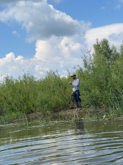A man stands on the muddy bank of a body of water, holding a fishing rod. He is wearing a gray hooded sweatshirt, blue jeans, a dark baseball cap, and green rubber boots. A dark fishing vest is slung across his chest. Behind him, dense green foliage lines the bank, and a line of trees extends into the background. The sky above is blue with scattered white clouds. The foreground shows the rippling surface of the water, reflecting the sky and surrounding vegetation. The location is identified as being in the city of Nezhinka, Russia.