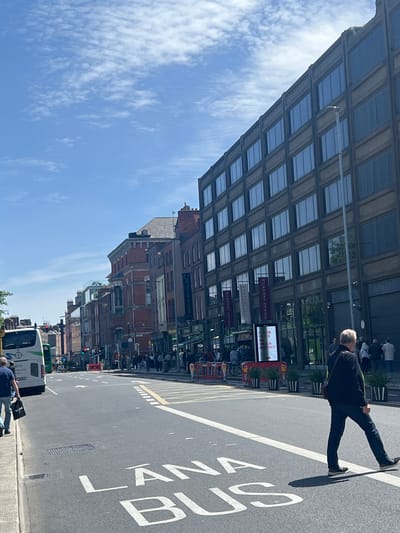 This is a vertical, eye-level, medium-angle shot of a street scene in Dublin, Ireland. The image captures a wide street with a bus lane marked with white paint that reads "LANA BUS". A white bus is parked on the left side of the street. People are walking on the sidewalk and crossing the street.
On the right side of the street is a large, multi-story building with many windows. The building has a modern design with dark-colored facade. Banners with "KILKENNY DESIGN" are hanging from the building. A man wearing a black jacket and blue jeans is crossing the street from right to left.
On the left side of the street, there are several older buildings made of red brick with traditional architecture. The sky is blue with scattered white clouds. The overall lighting suggests it is daytime with clear weather.