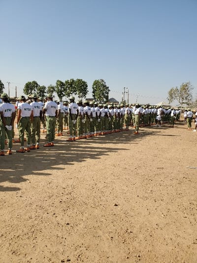 This image captures a large group of people, likely young adults, standing in organized rows on what appears to be an open dirt or sand field under a clear blue sky. The individuals are dressed in a uniform consisting of white t-shirts and what appear to be green trousers or skirts, and some are wearing caps. Their backs are mostly towards the camera, but enough of their torsos are visible to show text on their white t-shirts. The most prominent text visible is "NYSC", often accompanied by what looks like "NYSC D" or "D 03" or other letters/numbers, suggesting an affiliation with the National Youth Service Corps.
The people are lined up in a disciplined formation, stretching into the distance towards the left side of the frame. Long, distinct shadows are cast from each person, extending across the dusty ground towards the right side of the image, indicating that the sun is relatively low in the sky, suggesting either early morning or late afternoon. The ground is a light tan or brown color, characteristic of dry earth, with small specks or stones scattered across it. In the background, beyond the formation, there are scattered green trees and what appear to be utility poles or structures. The overall impression is of a formal outdoor gathering, possibly a drill, parade, or assembly. The weather is bright and sunny.