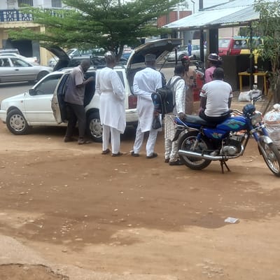 The scene takes place in Dadi Riba, Nigeria. Several men, some wearing white traditional attire, are standing near a row of white cars with open doors and trunks. One man sits on a blue motorcycle. The ground is dirt. Buildings are visible in the background.