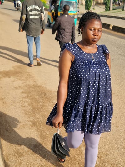 The image shows a street scene in the city of Kabong, Nigeria. In the foreground, a woman walks towards the viewer, carrying a black handbag. She is wearing a dark blue dress with white polka dots and light purple tights. Behind her, a man in a dark jacket and blue jeans walks away from the camera. Further in the background, a small, three-wheeled vehicle, possibly a tuk-tuk, is visible. The ground is a sandy, dirt road. The lighting suggests it is daytime, with shadows cast on the ground, indicating the sun is at an angle. The environment appears to be an urban or semi-urban area with some vegetation and buildings lining the street.