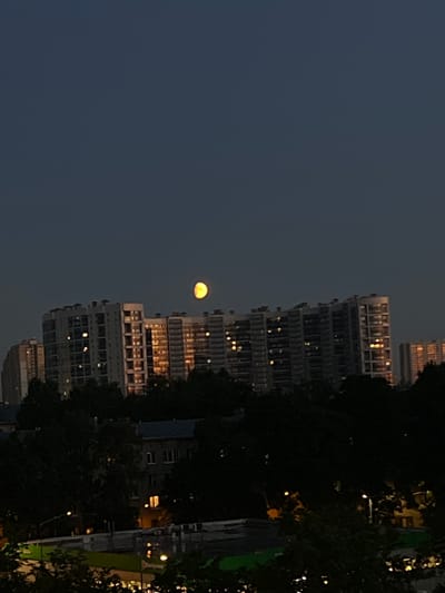 A full moon, illuminated by an unseen light source, hovers in a dark twilight sky above a cityscape. The moon, a bright yellow orb, is partially obscured by a cluster of tall, modern buildings. These buildings are characterized by numerous windows, many of which are lit, suggesting occupancy. In the foreground, darker, lower-rise buildings and dense foliage create a stark contrast against the illuminated structures and the moon. The overall scene depicts an urban environment during dusk or early night.