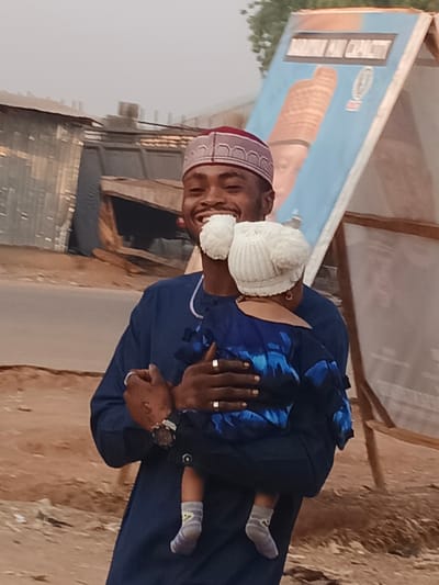 An adult male with dark skin, wearing a navy blue long-sleeved tunic with white trim and a maroon and light-colored patterned cap, is depicted holding an infant/toddler. He smiles broadly, revealing teeth, and wears two silver-colored rings on his left hand and a dark-faced watch on his left wrist. The child, held in the male's left arm and facing away from the camera, wears a white knitted hat with two pom-poms, a blue patterned top, dark trousers, and grey socks with a yellow design.
The scene is set outdoors on a street in Doma, Nigeria. In the background to the left, multiple structures made of corrugated metal sheets are visible. To the right, a large blue signboard with a depiction of a person wearing a similar traditional cap and partial text, including "MAN CAPACITY," stands on wooden supports. The foreground consists of light brown, dusty soil, transitioning to a darker, possibly paved, surface behind the subjects. The sky is hazy, and minimal green foliage is visible at the upper edge of the frame.