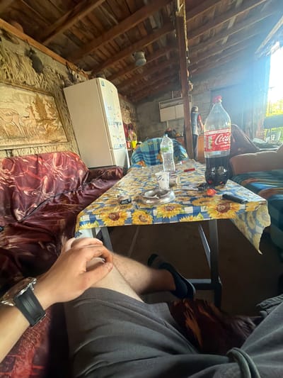 The photo depicts an interior scene in a rustic setting, possibly Krivo Pole, Bulgaria, given the prompt. The location appears to be an attic space, as evidenced by the exposed wooden beams of the roof structure. Natural light floods the area from an unseen source.
A person is seated at a table covered with a blue and yellow sunflower-patterned tablecloth. The table holds various items, including a large Coca-Cola bottle, a water bottle, an ashtray, and what appear to be cigarettes. The person's wrist, adorned with a large wristwatch, is visible.
The seating arrangement consists of a chair covered in a maroon-patterned fabric. Adjacent to the chair is a white appliance, likely a refrigerator or freezer. The floor is covered in a textured material that is partly obscured.